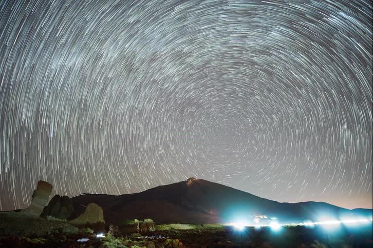 A breathtaking timelapse captures circular star trails rotating in the night sky over a volcanic mountain landscape