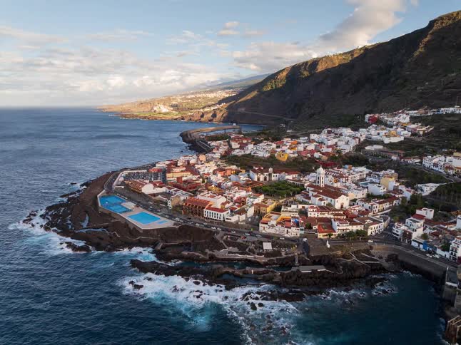 Sweeping aerial reveal of the picturesque coastal town of Garachico in Tenerife, showcasing its natural swimming pools and the iconic Mount Teide in the distance