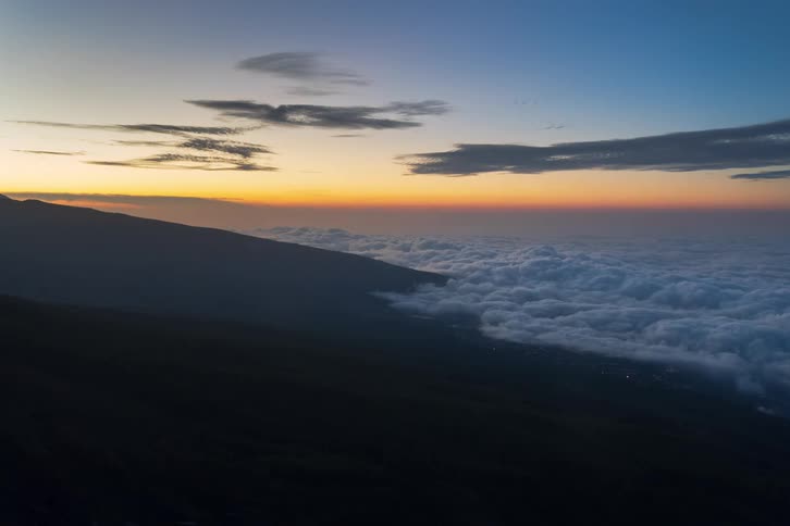 High-altitude timelapse of a colorful sunset fading to night over a vast sea of clouds, revealing a glowing city beneath