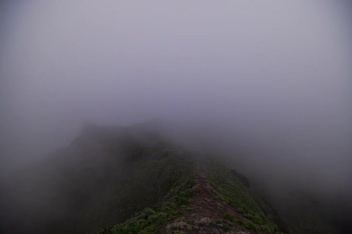 A dramatic time-lapse shows thick fog swallowing a green mountain ridge as hikers navigate the misty trail