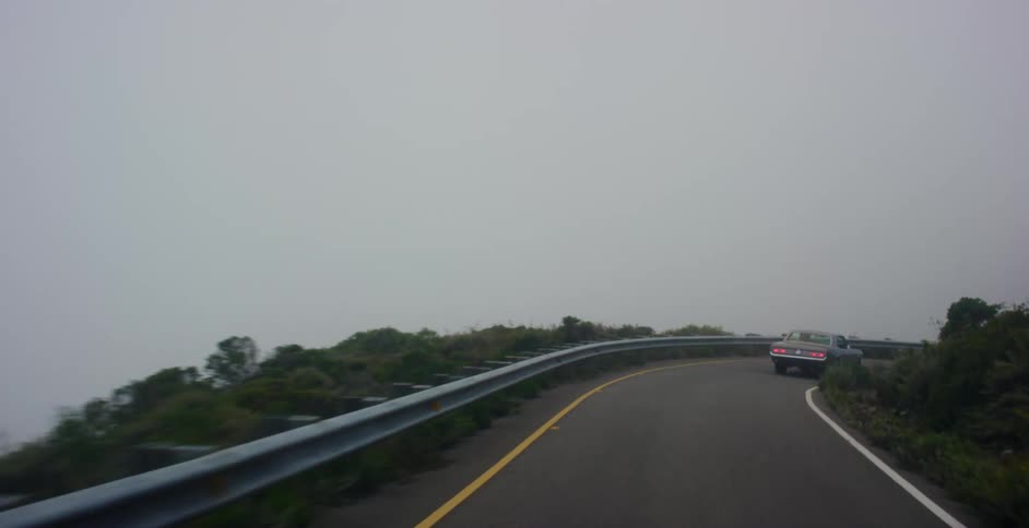 Following a classic American muscle car as it drives along a winding mountain road shrouded in dense fog