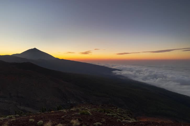 A stunning day-to-night timelapse shows the sun setting behind a majestic mountain peak, casting long shadows over a sea of clouds before revealing city lights below