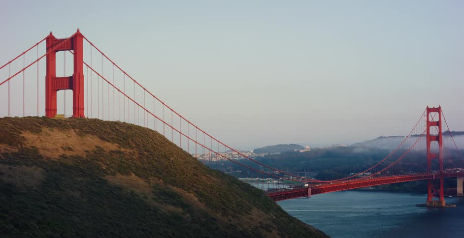 Static wide shot of the Golden Gate Bridge partially obscured by a hill at sunrise, with traffic crossing and a boat on the water