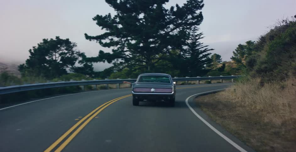 POV following a classic dark muscle car as it drives along a scenic, winding road through a misty, hilly landscape