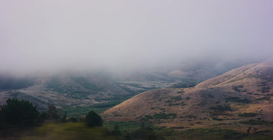 A moody tracking shot reveals a vast valley of rolling hills partially obscured by a thick layer of fog