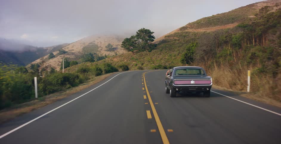 A person hangs their arm out the window of a classic Ford Mustang as it drives along a scenic, winding country road through foggy hills