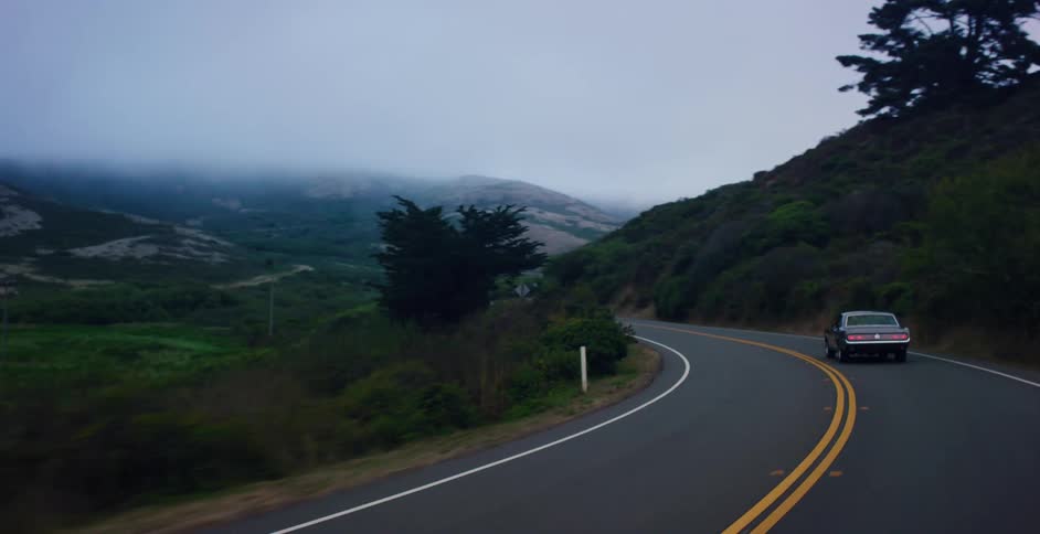 A classic American muscle car drives along a winding coastal highway through foggy, green hills