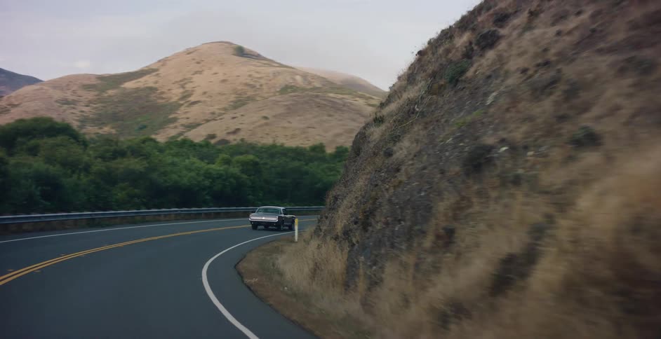 A point-of-view perspective from a car following a vintage pickup truck along a winding rural highway through rolling hills