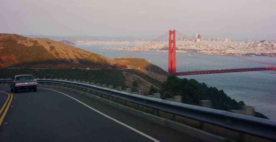A classic car drives along a winding scenic road overlooking the Golden Gate Bridge and the San Francisco skyline at sunrise