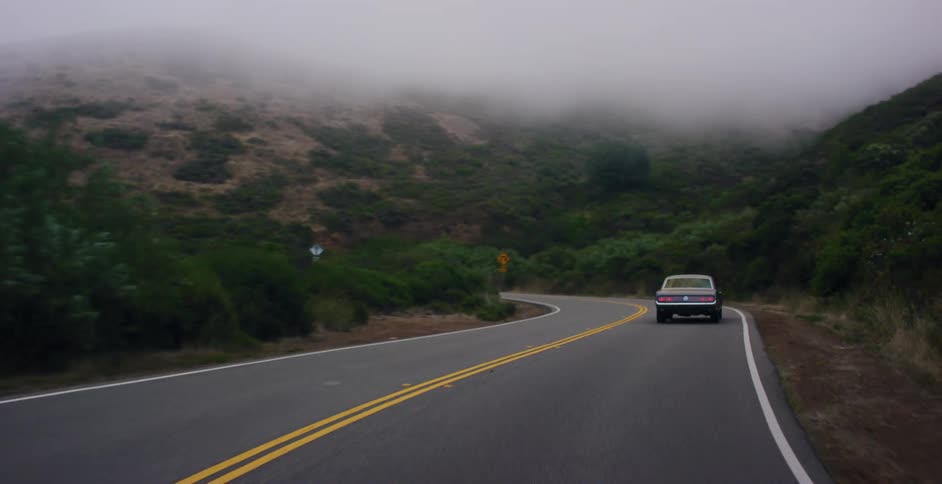 A classic car drives along a winding road into the foggy, green hills