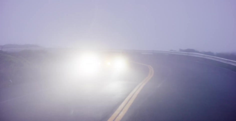 A classic muscle car emerges from the dense fog, its bright headlights cutting through the gloom as it drives around a winding road