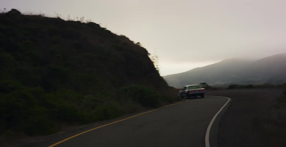 A classic car drives away from the camera and around a sharp curve on a foggy, misty coastal road