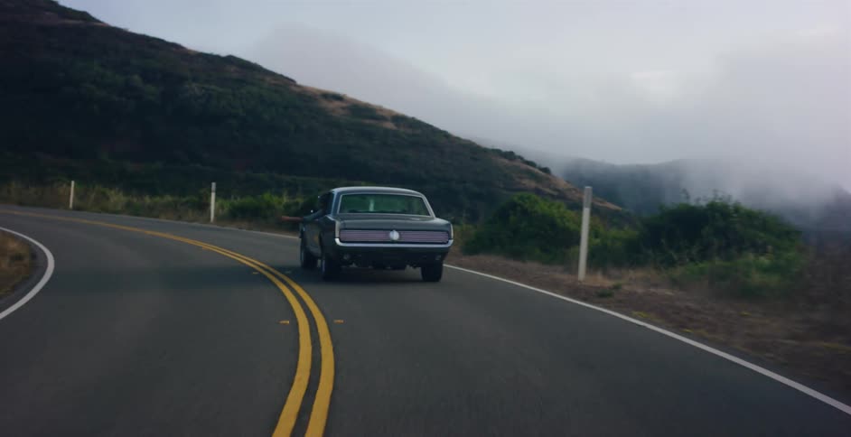 A classic Ford Mustang cruises down a winding coastal highway, navigating through the misty mountain air on a scenic road trip