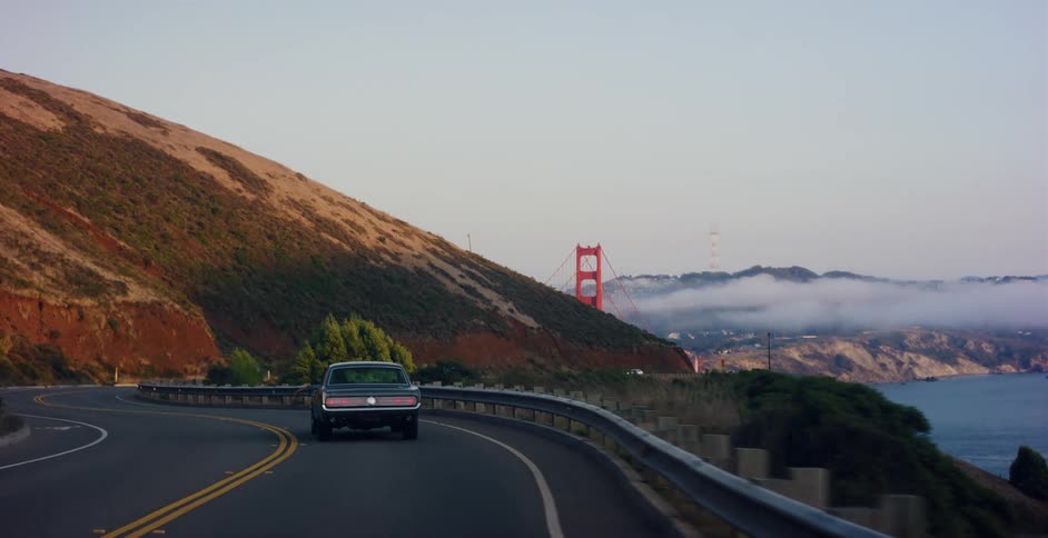 A classic muscle car cruises along a winding coastal highway with the Golden Gate Bridge visible in the distance at sunset