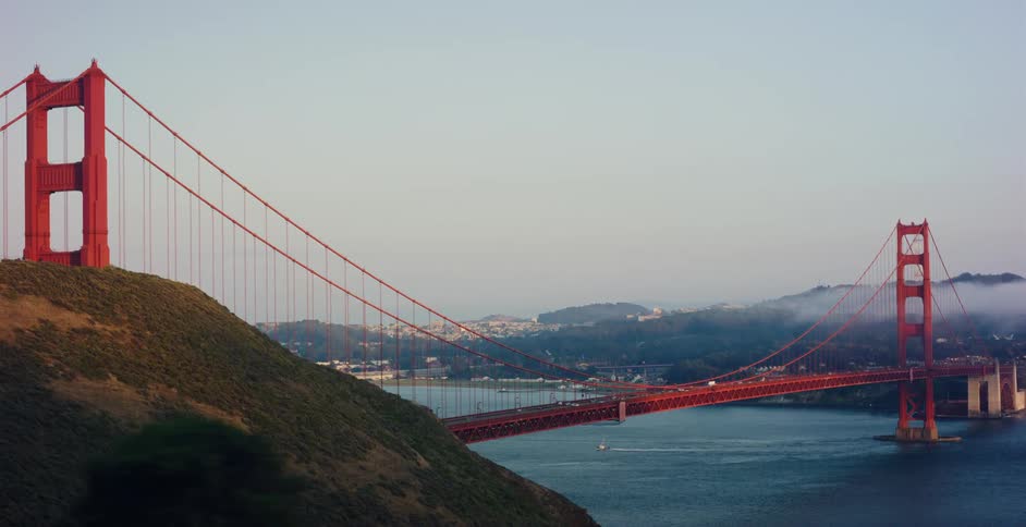 Iconic wide shot of the Golden Gate Bridge in San Francisco as traffic flows across and a sailboat passes below at sunrise