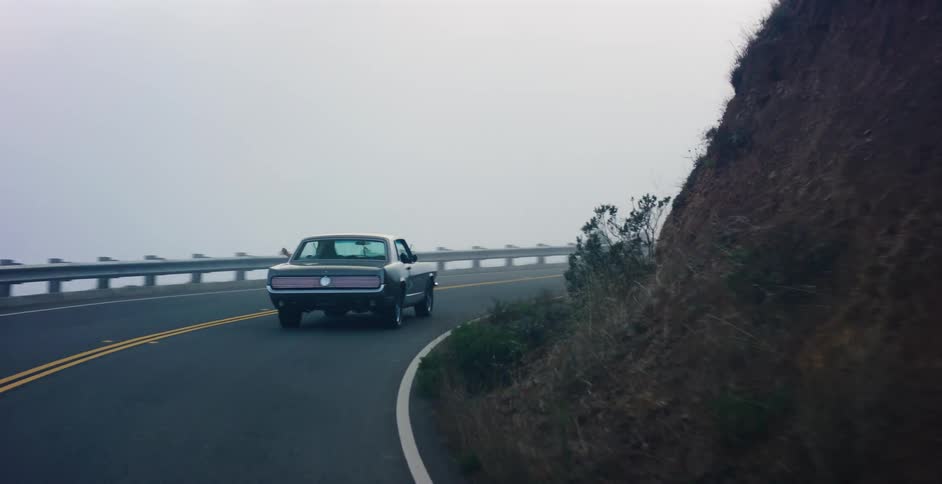 Rear view of a classic muscle car driving along a winding coastal highway into the dense fog