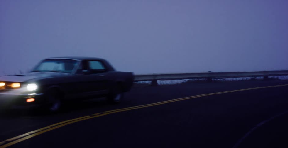 A classic black Ford Mustang with its headlights on drives slowly around a bend on a dark, foggy mountain road