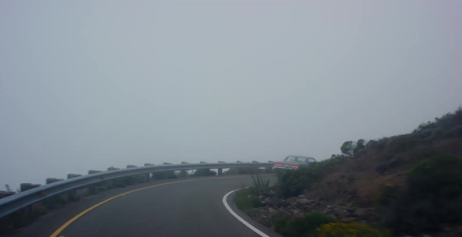 A point-of-view shot follows a classic American muscle car as it drives along a foggy, winding coastal highway