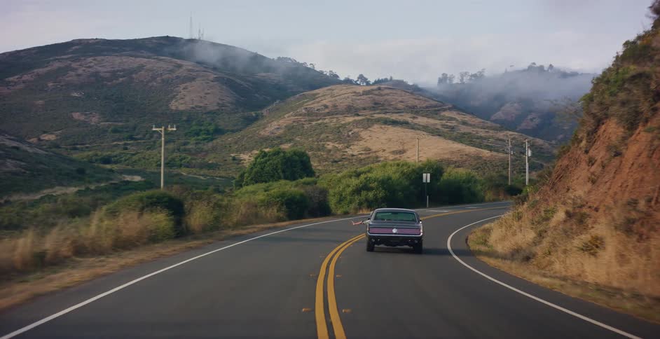 A classic muscle car travels down a winding road through a lush, foggy mountain landscape on a scenic road trip