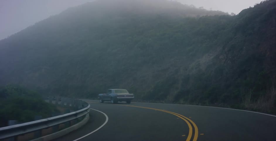 A vintage muscle car navigates a winding coastal highway enveloped in thick, moody fog