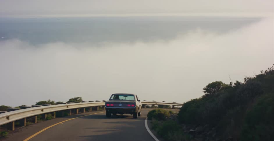 A classic Mustang cruises along a stunning coastal highway overlooking a city before driving into a dense bank of fog