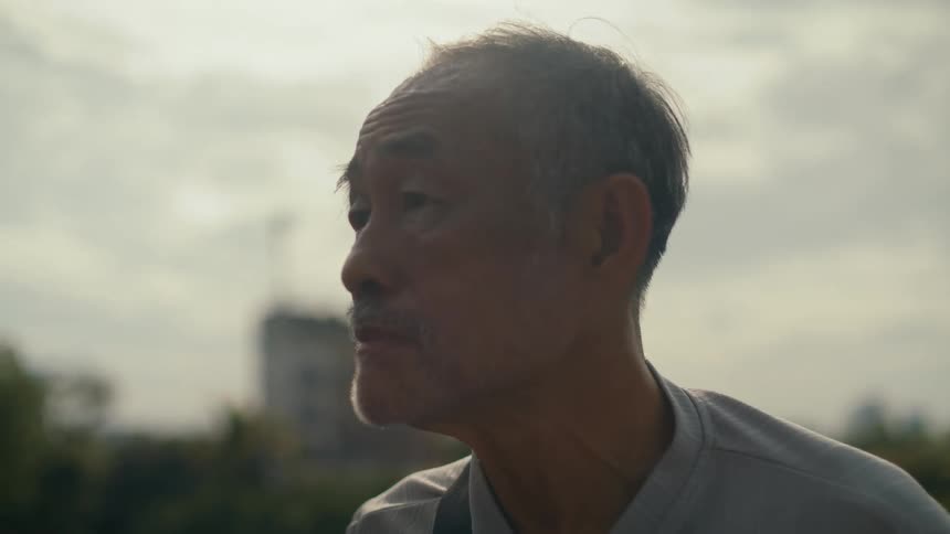 A contemplative elderly Asian man gazes into the distance with a city skyline in the background as the camera slowly focuses on his face