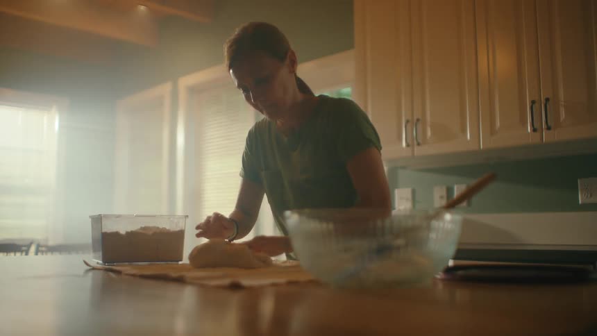 A woman kneads bread dough on her kitchen counter as warm, hazy sunlight streams in through the window