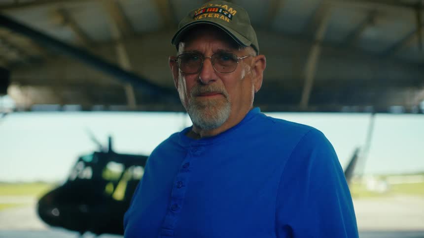 A proud Vietnam War veteran stands stoically in an airplane hangar with a helicopter in the background