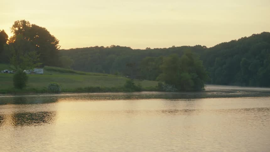 A tracking shot from a moving vehicle reveals a tranquil lake at sunrise, with golden light reflecting on the water and forested hills in the background