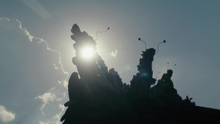 The sun peeks out from behind the silhouette of an ornate Asian temple roof against a cloudy sky