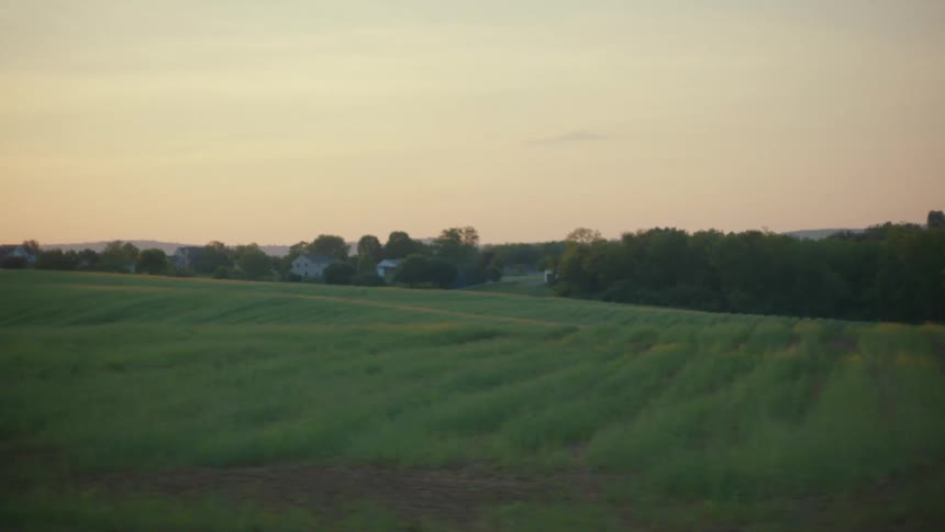 A tranquil view of a rolling green countryside field with distant homes seen from a moving vehicle at sunset