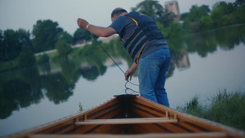 From an interior perspective, a man wearing a life jacket readies a wooden canoe's rope for launch on a tranquil lake at dusk