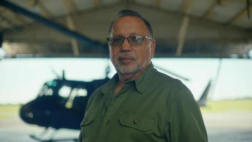 A stoic portrait of an older man standing in an aircraft hangar with a helicopter in the background