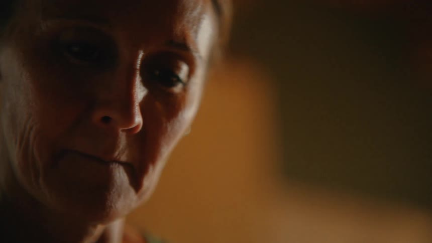 A slow-motion close-up captures the worried and somber expression of an elderly woman in a dimly lit, warm environment
