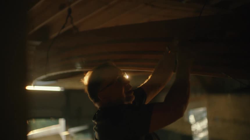 A dedicated craftsman sands a wooden canoe suspended from the ceiling of his backlit workshop