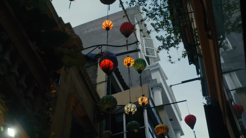 Illuminated colorful lanterns hang across a narrow city alleyway against the darkening sky