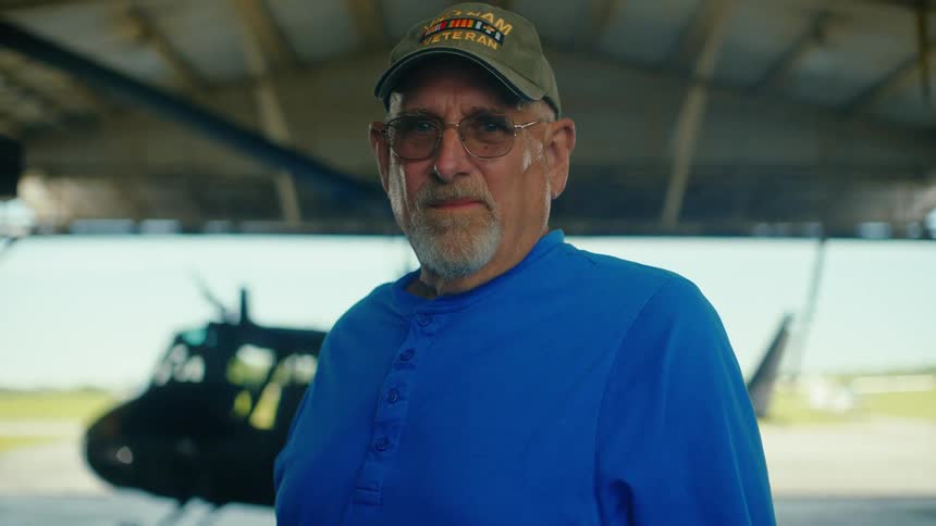 Portrait of a Vietnam veteran wearing a cap and glasses, standing seriously in a hangar with a helicopter in the background