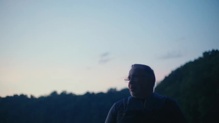 A contemplative older man wearing glasses is silhouetted against the tranquil twilight sky in nature