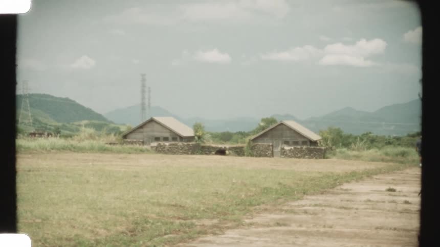 A static shot from vintage 8mm film shows two rustic barns sitting in a grassy field with rolling green hills in the background