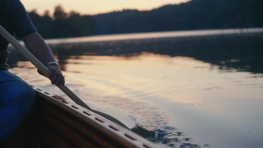 A person paddles a wooden canoe across a calm lake at sunset in serene slow motion