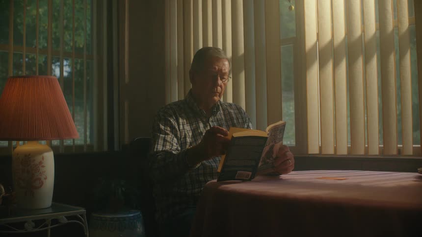 An older man peacefully reads a book while sitting at a table as warm sunlight filters through the window blinds