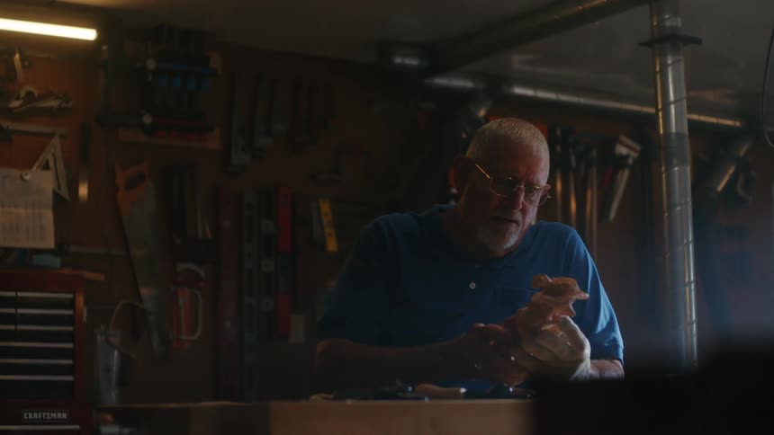 A senior woodworker carefully examines a wooden carving at his workbench in a dimly lit workshop