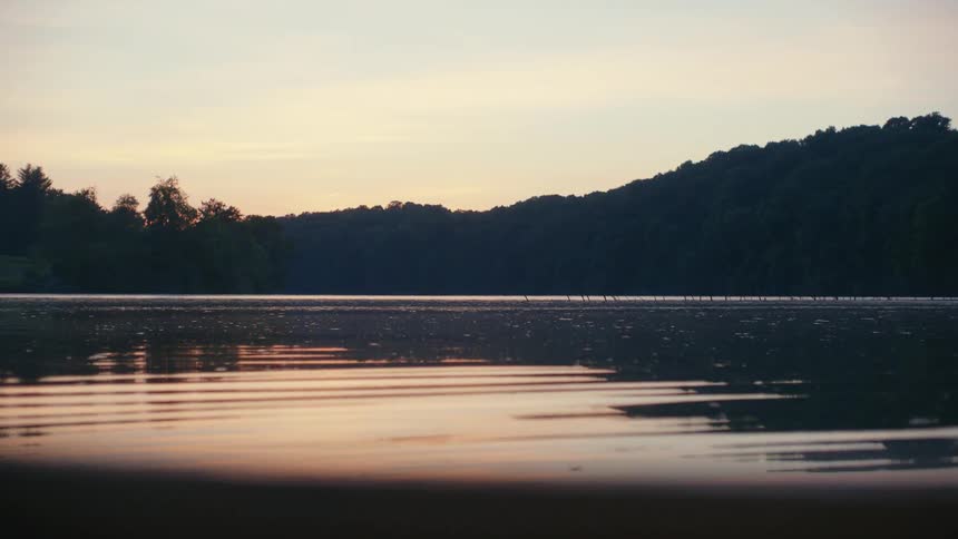 A tranquil lake gently ripples, reflecting the soft, warm light of an early morning sunrise over a forested hill