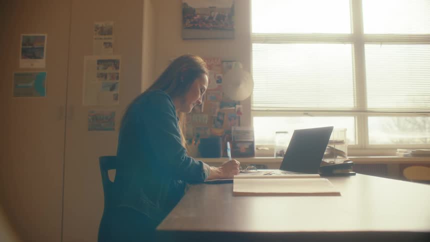A woman smiles as she works at her desk, writing on paper next to her laptop in a sunlit office