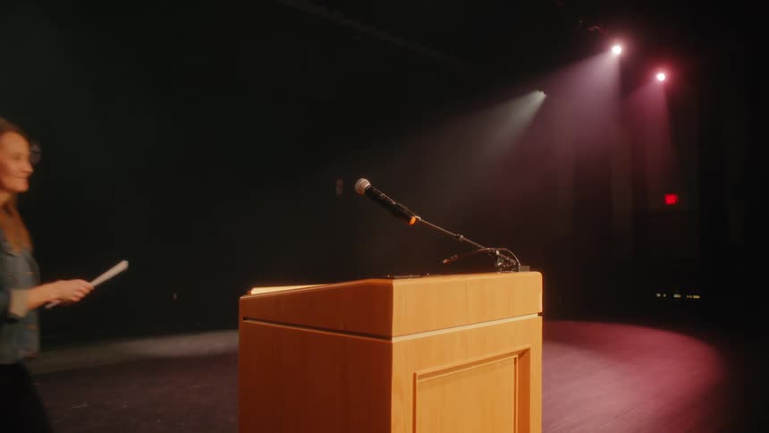 A smiling woman walks to a podium on a dark stage to give a presentation under a single spotlight