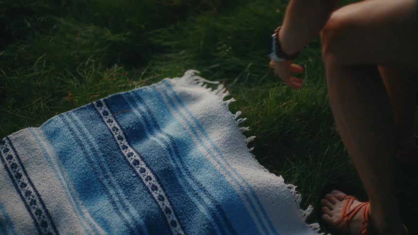 A woman carefully lays out a blue and white striped blanket on a green lawn for a picnic in slow motion