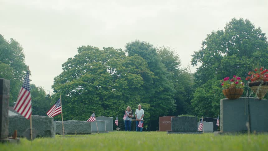 An elderly couple pays their respects at a loved one's grave in a cemetery decorated with American flags
