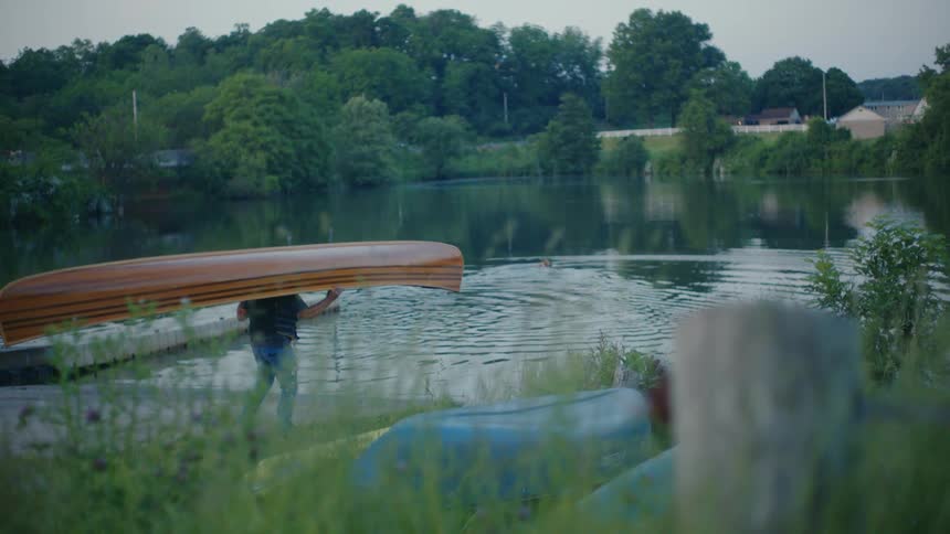A person portages a wooden canoe along a grassy bank towards a calm lake at dusk