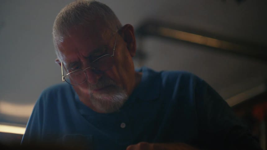 A close-up of an elderly man with glasses and a grey goatee concentrating on a task in his dimly lit workshop
