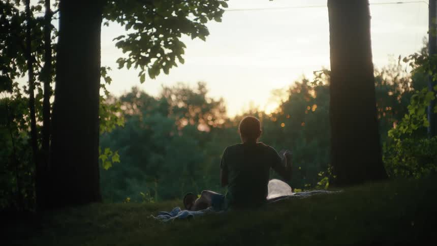 A silhouetted couple enjoys a peaceful picnic on a grassy hill framed by two trees at sunset